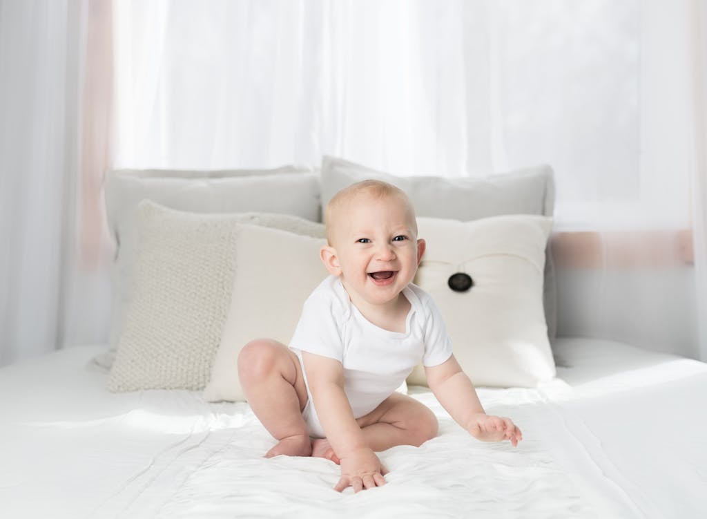 Cute baby sitting and smiling on a white bed in a bright, airy bedroom.